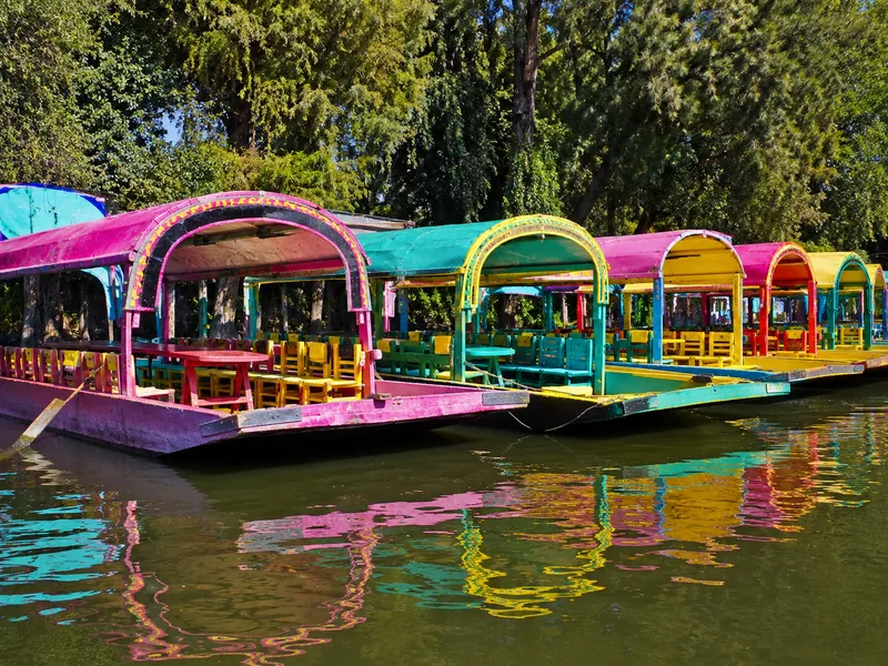 Trajineras in einer Reihe vertäut, von der Wasserlinie aus gesehen, mit bogenförmigen Dächern in Pink, Türkis und Gelb über grünem Kanalwasser